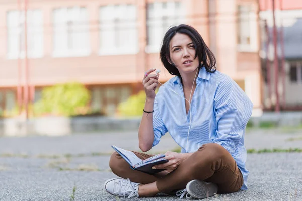 Disturbed European beautiful woman while sitting at sidewalk, studying and making notice on notebook  planner. Young woman with earphone listen and preparing for exam.