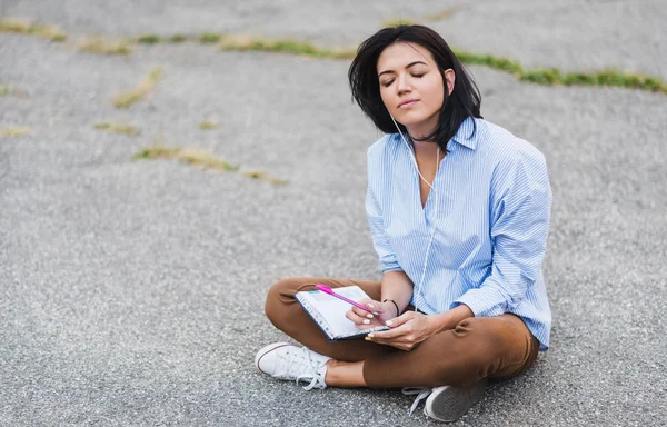 Казуальная стильная кавказская студентка, сидящая на тротуаре, наслаждаясь онлайн-общением с помощью беспроводного доступа в Интернет на своем смартфоне. Молодая женщина с наушниками слушать и готовиться к экзамену . — стоковое фото