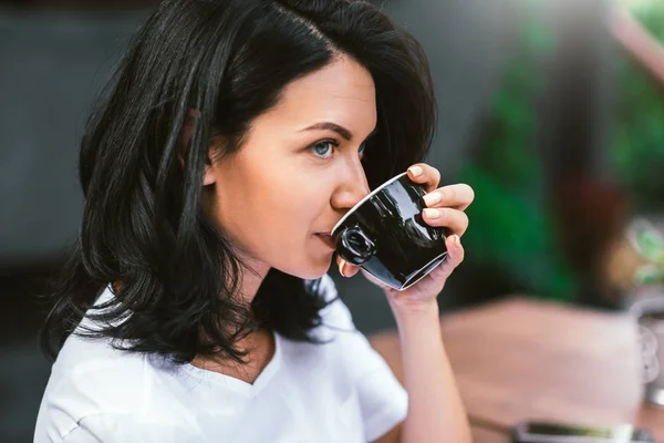 Jolie fille brune caucasienne vêtue de t-shirt blanc buvant du café, détournant les yeux avec une expression lourde et sérieuse, faisant des plans pour la journée. Concept de personnes et mode de vie . — Photo