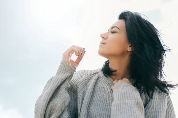 Vista lateral da morena caucasiana jovem viajante com cabelo ventoso, sonhando contra o céu luz solar. Mulher sonhadora com olhos fechados em pulôver cinza. Estilo de vida e conceito de pessoas. Humor de cobertura . — Fotografia de Stock