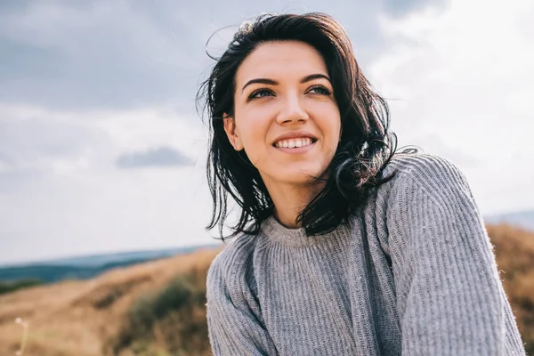 Retrato de mulher morena feliz e engraçada sorrindo e olhando para o lado, contra o prado da natureza e fundo nublado com cabelo ventoso. Pessoas, viagens e conceito de estilo de vida. Humor de cobertura . — Fotografia de Stock