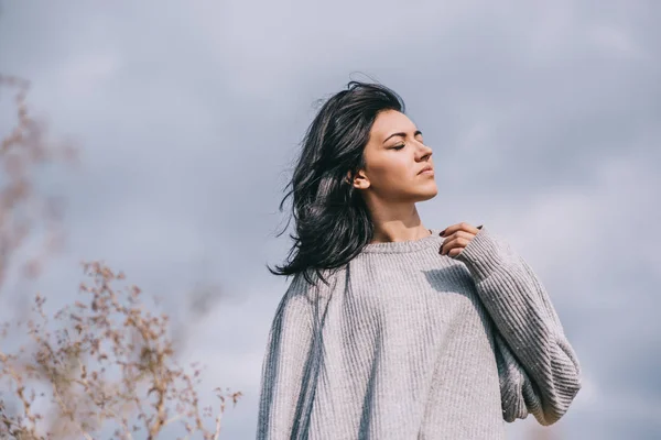 Visão inferior de atraente morena caucasiana jovem viajante com cabelo ventoso, posando contra o céu nublado. Mulher sonhadora com olhos fechados em pulôver cinza. Estilo de vida e pessoas conceito humor . — Fotografia de Stock