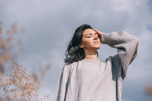 Lifestyle and people concept mood. Bottom view of attractive brunette Caucasian young woman traveler with windy hair, posing against overcast sky. Dreamy female with closed eyes in gray pullover. — Stock Photo, Image