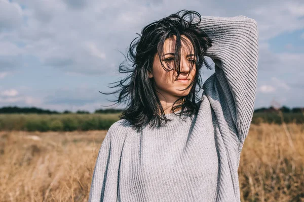 Morena caucasiana jovem com cabelo ventoso soprando fora, contra prado e céu. Retrato de mulher de sonho olhando para longe com a mão na cabeça. Estilo de vida conceito de moda. Ideia de capa humor . — Fotografia de Stock