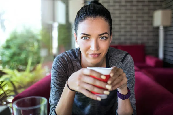 Indoor tiro de atraente alegre jovem mulher caucasiana com xícara de café sentado no restaurante, olhando para a frente da câmera durante o café da manhã . — Fotografia de Stock