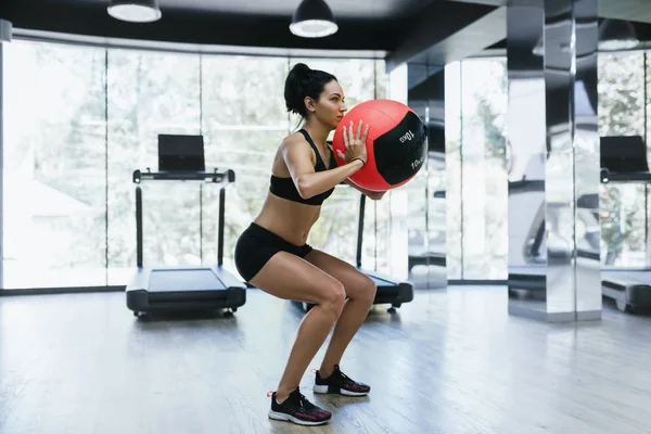 Mujer atlética activa haciendo ejercicios de sentadillas en el gimnasio. Fuerte sentadilla femenina en la alfombra de fitness con balón de medicina de peso en el club de salud. Deporte, personas y estilo de vida . — Foto de Stock