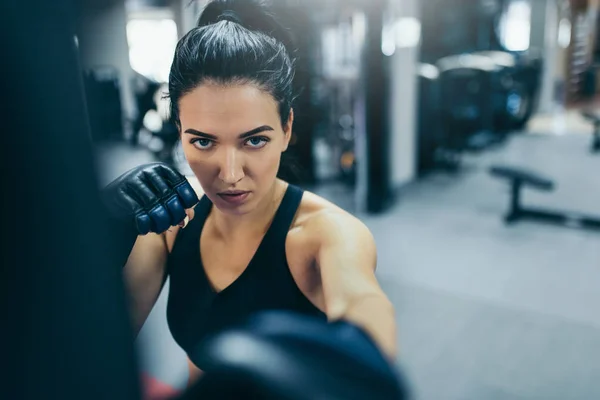 Atractiva entrenadora morena golpeando una bolsa con guantes de kickboxing en el gimnasio y mirando al frente a la cámara. Concepto de deporte, fitness, estilo de vida y motivación . — Foto de Stock