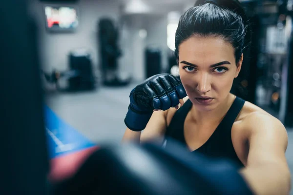 Front view of strong brunette attractive female trainer punching a bag with kickboxing gloves in the gym workout. Sport, fitness, lifestyle and motivation concept. — Stock Photo, Image
