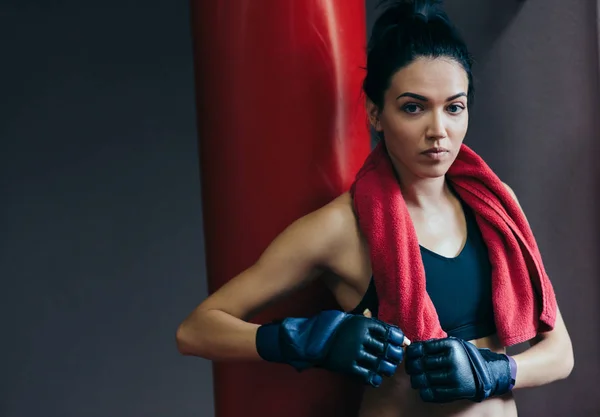 Horizontal portrait of strong attractive brunette woman with red towel on neck before workout with punch bag, wearing kickboxing gloves in the gym. Sport, fitness, lifestyle and people concept. — Stock Photo, Image