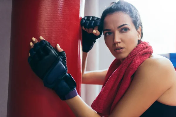 Retrato de cerca de la atractiva mujer morena deportiva con toalla roja en el cuello después del entrenamiento en saco de boxeo rojo, usando guantes de kickboxing en el gimnasio. Deporte, fitness, estilo de vida, personas, concepto de motivación . — Foto de Stock