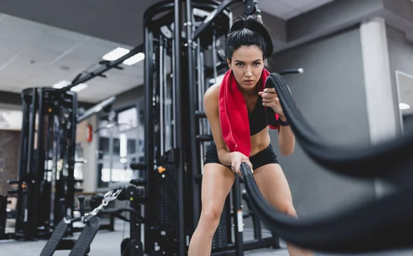 Sport, people, motivation concept. Fitness active brunette sportswoman working out in functional training gym doing crossfit exercise with battle ropes with red towel on neck, copyspace for your text — Stock Photo, Image