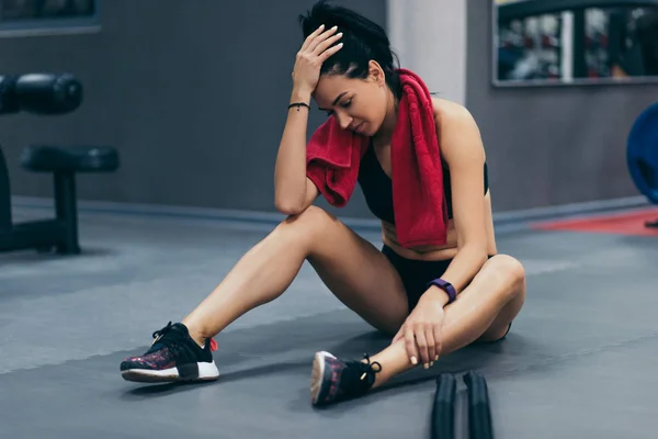 Young brunette sportswoman sitting on a floor at gym after her battle rope workout and looking down. Attractive female athlete taking rest after fitness training at gym with red towel on neck. — Stock Photo, Image
