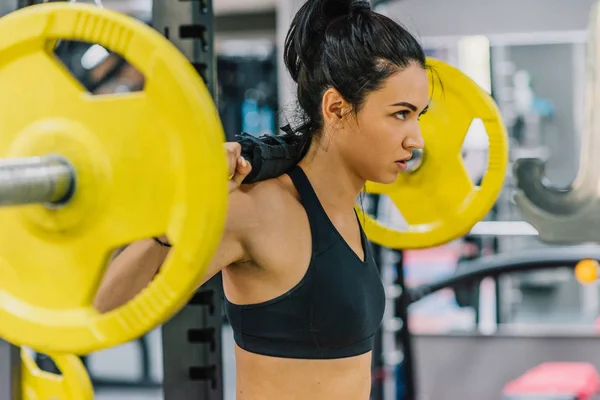 Side view of beautiful young woman doing workout in the gym, pushing yellow barbell on shoulders. Female hard workout for cross fit. People, sport, fitness, healthy lifestyle and motivation concept. — Stock Photo, Image