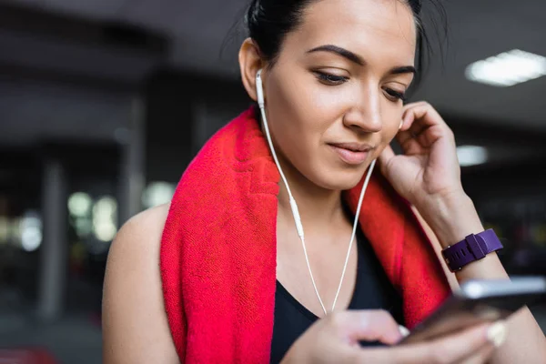 Retrato recortado de uma bela jovem sorridente e esportiva ouvindo música de seu smartphone, tendo uma pausa após o treino no ginásio. Pessoas, tecnologia, fitness e conceito de esporte . — Fotografia de Stock