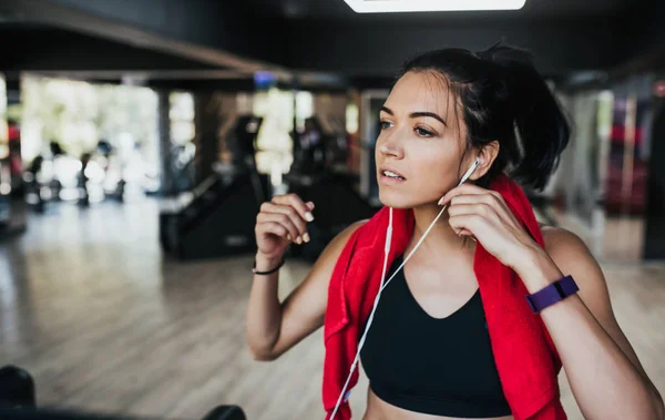 Retrato horizontal joven mujer caucásica corredor corrige los auriculares mientras corre en el gimnasio, usando ropa deportiva negro y toalla roja en el cuello. Concepto de personas, deporte y fitness . — Foto de Stock
