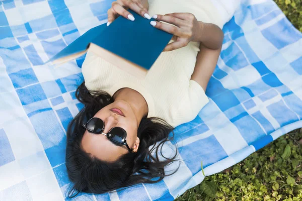 Genç Beyaz esmer bir kadın Parkta piknik sırasında bir kitapla yeşil çim üzerinde yalan samimi bir kadeh. Güzel kız öğrenci sunglasess giyen ve açık bir kitap okuma. Yaşam tarzı, eğitim. — Stok fotoğraf