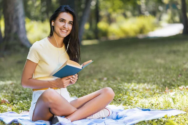 Повне тіло подання кавказьких брюнетка красиві молоді усміхається жінка з навушники, слухати музику і читати книгу в парку. Привабливі щасливі жіноча розслаблюючій в парку під час пікнік. Копія простір — стокове фото