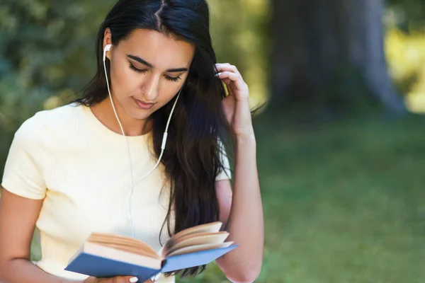 Müzik ve parkta bir kitap okuma güzel beyaz esmer genç bir kadın ile kulaklık, kırpılmış bir kadeh. Çekici kadın styding ve kitap üzerine ders öğrenme. Kopya alanı — Stok fotoğraf
