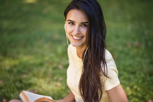 Candid portrait of Caucasian brunette beautiful young smiling woman with earphones, listening to music and reading a book at park. Attractive happy female relaxing in the park during picnic.Copy space