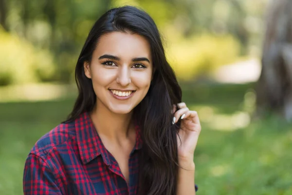 Attrayant plan de heureuse jeune brune caucasienne souriante et portant une chemise à carreaux sur fond nature dans le parc. Style de vie, personnes et concept de beauté . — Photo