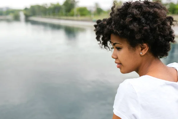 Vue latérale du beau jeune modèle noir heureux avec une coiffure bouclée courte regardant vers le lac dans le parc, posant à l'extérieur. Style de vie, émotions et personnes concept de style de vie . — Photo