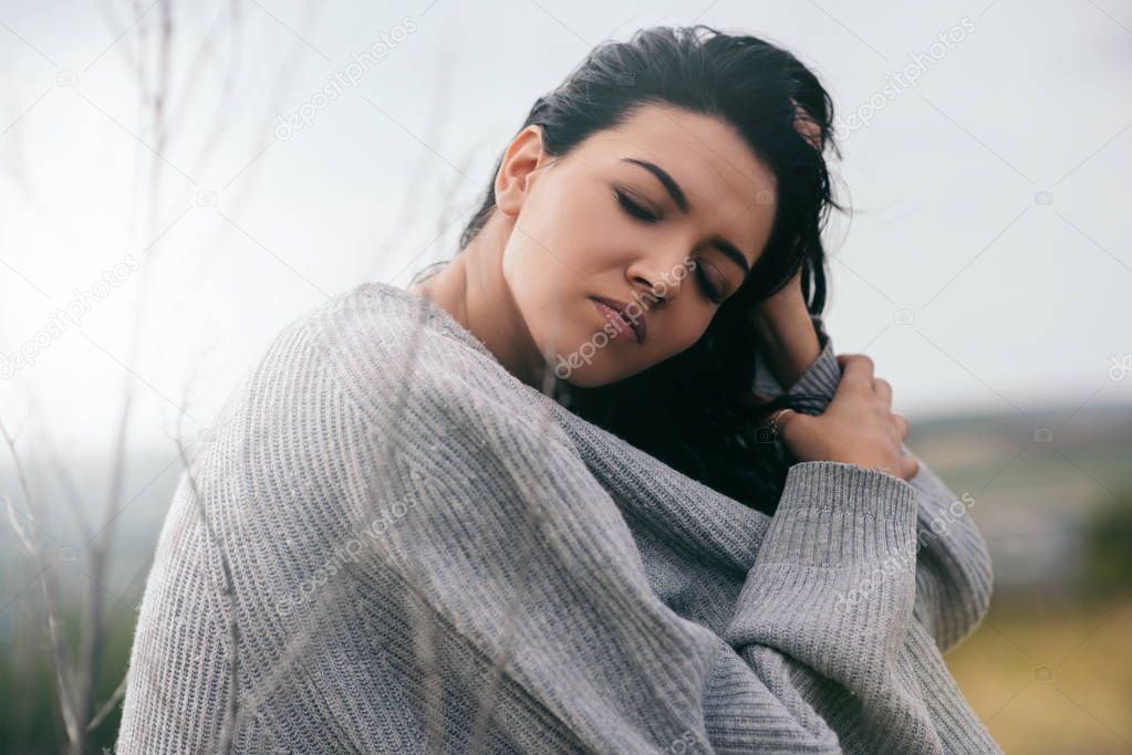 Portrait of attractive brunette caucasian young woman traveler with windy hair, dreaming outside in the park. Dreamy female with closed eyes in gray pullover. Lifestyle and people concept. Cover mood.