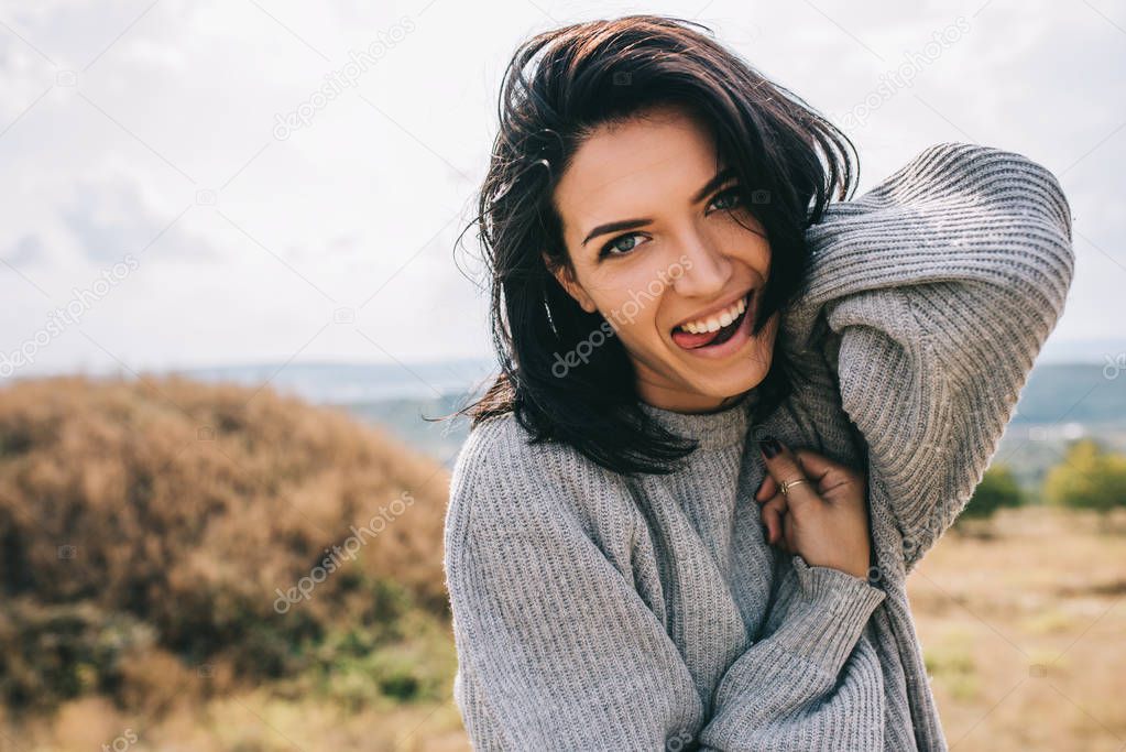 Happy and funny brunette woman smiling and stick out tongue against nature meadow background with windy hair, having fun during her vacation in mountain. People, travel, lifestyle concept. Cover mood