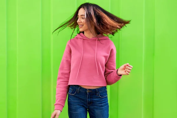행복 한 십 대 소녀가 분홍색 후드와 청바지를 입고 활짝 웃으며 머리를 흔들며 춤을 추고 있습니다. 아름다운 암컷의 옥외 모습은 푸른 배경 위에서 즐거운 표현을 한다. — 스톡 사진