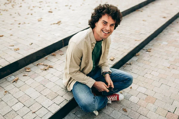 Handsome young man standing outdoors, typing messages on mobile phone. Young male with curly hair — Stock Photo, Image