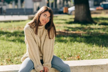 Kameraya bakan güzel, gülümseyen bir öğrenci kadın resmi. Şehir caddesinde oturuyor. Örgü süveter ve gözlük takıyor. Güneş ışığında poz veren güzel genç bir dişi.