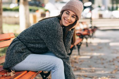 Kameraya bakan güzel, gülümseyen bir kız öğrencinin görüntüsü, şehir caddesinde bankta oturuyor, örgü örülmüş süveter, gözlük ve şapka giyiyor. Güzel genç bayan dışarıda dinleniyor.