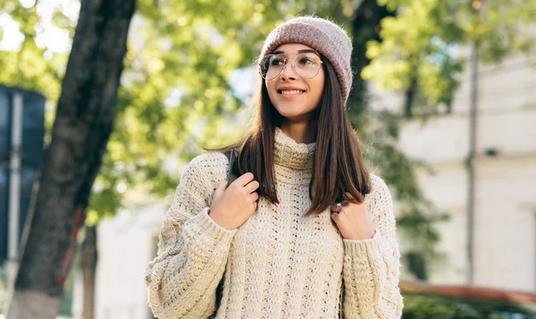 Utendørsbilde av smilende studentkvinne som ser til side, går på gaten om morgenen, i strikket genser, briller og hatt. Vakker ung kvinne med ryggsekk som poserer utenfor – stockfoto