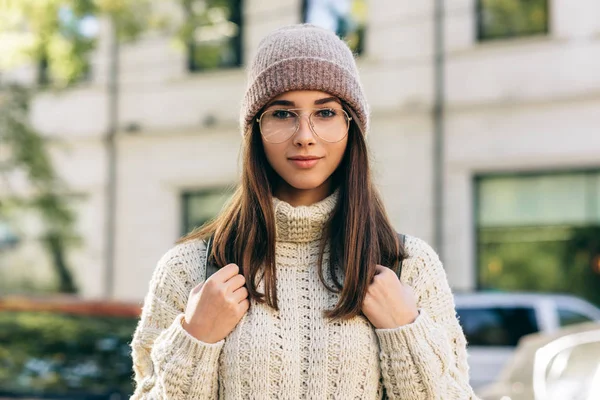 Retrato al aire libre de una estudiante sonriente mirando a la cámara, de pie en la calle por la mañana, con suéter de punto, anteojos y sombrero. Hermosa joven con mochila posando fuera —  Fotos de Stock