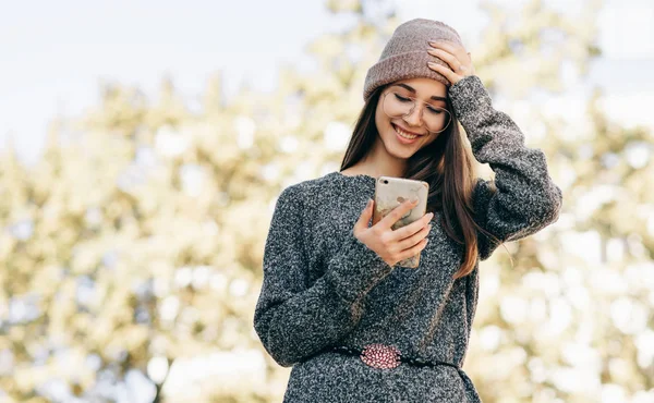 Imagem de uma jovem sorridente em pé ao ar livre conversando com seu amigo no telefone inteligente. Mulher morena vestida com camisola de malha, chapéu, óculos navegando online na rua da cidade . — Fotografia de Stock