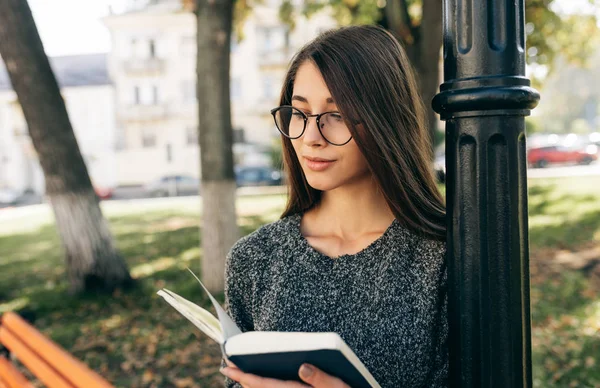 Досить молода жінка в светрі і прозорих окулярах, стоячи на вулиці міста і читаючи книгу, чекаючи на своїх друзів. Молода студентка навчається на міській вулиці . — стокове фото