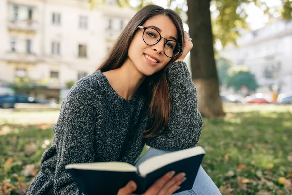 Bild av vackra leende ung kvinna bär tröja och transparenta glasögon, sitter utomhus iin stadsparken, läsa boken. Unga kvinnliga student lärande i staden gatan. — Stockfoto