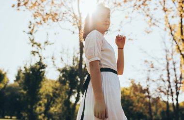 Beyaz elbiseli beyaz bir kadın güneş ışığı ve doğa arka planında poz veriyor. Güneşli bir günde parkta yürürken gülümseyen güzel bir kadın..