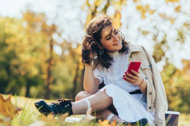 Neşeli genç bir kadının elinde tek kullanımlık bir fincan kahve, parkta yürürken telefonla fotoğraf çekmesi. Güzel öğrenci hazır kahve içiyor ve internetten cep telefonuna bakıyor.. 