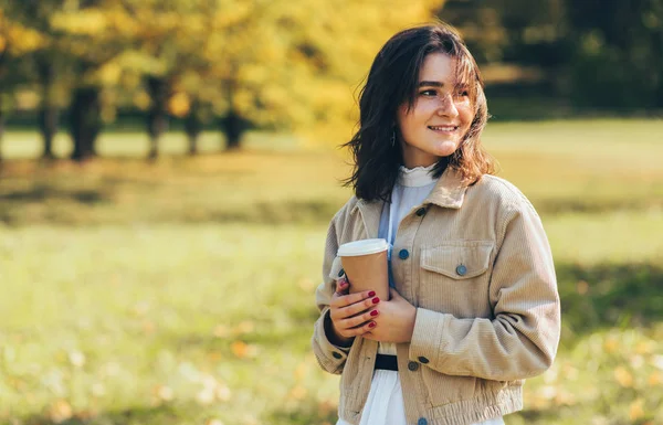 Sorrindo bela jovem mulher vestindo suéter e jaqueta e segurando xícara descartável de café no parque da cidade. Estudante bonita bebendo café takeaway ao ar livre pela manhã . — Fotografia de Stock