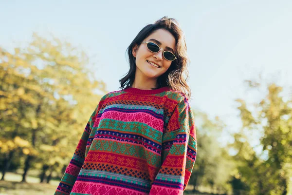 Feliz bela jovem mulher sorrindo amplamente com cabelo ventoso e sardas, vestindo suéter e óculos de sol posando na natureza fundo de luz solar no parque . — Fotografia de Stock