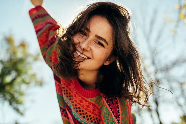 Visão inferior close-up retrato da jovem mulher feliz sorrindo amplamente com cabelo ventoso e sardas tem expressão alegre, vestindo camisola de malha colorida com os braços abertos, posando na luz solar da natureza — Fotografia de Stock