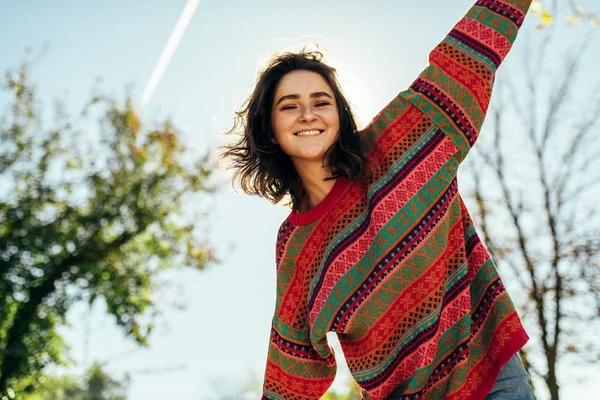 Visão inferior imagem de feliz bela jovem sorrindo amplamente com cabelo ventoso e sardas tem expressão alegre, vestindo camisola de malha colorida com os braços bem abertos, posando na luz solar da natureza — Fotografia de Stock