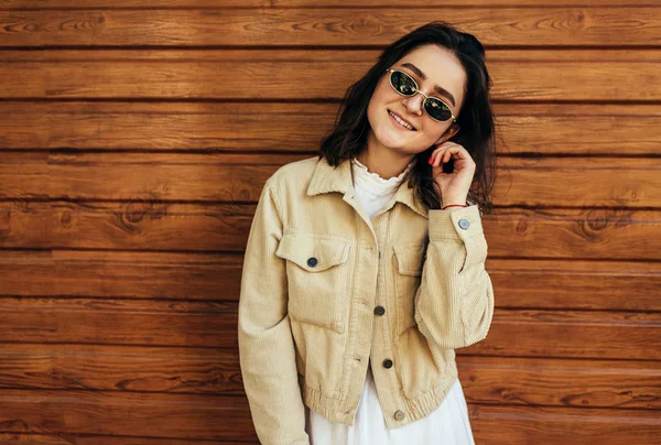 Imagem de beleza natural jovem mulher vestindo óculos de sol na moda, tem expressão alegre, posando na rua da cidade na parede de madeira. Estudante bonita fêmea tendo um descanso antes da adversidade . — Fotografia de Stock