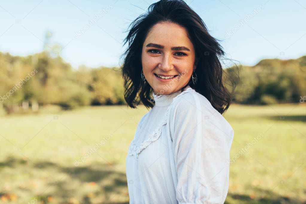 Beautiful smiling young woman with windy hair enjoying the warm weather, wearing white dress on nature background. Pretty natural beauty female smiling during walking in the park on sunny day.