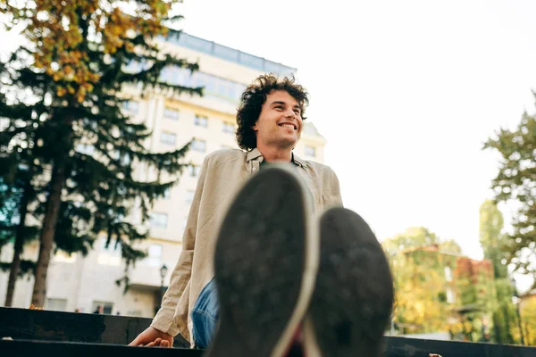 Bottom view image of positive handsome young man sitting on the stairs, smiling and looking at one side. Outdoor image of happy smart student male resting outside. People and emotion concept