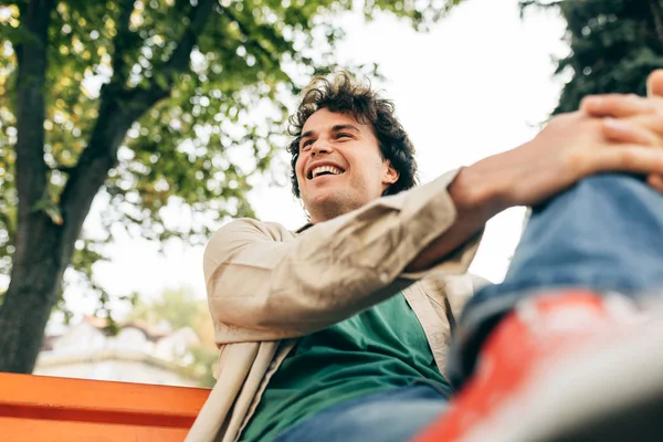 Bottom view of happy smiling man with curly hair sitting on the bench outdoors in the city street. Excited cheerful student male making good gesture.