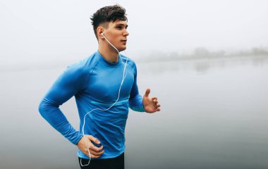 Sis gölü arka planında sabah koşan genç bir koşucunun yatay açık hava görüntüsü. Sporcu erkek parkta egzersiz yapıyor ve kulaklıkla müzik dinliyor. İnsanlar ve spor konsepti