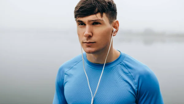 Horizontal close-up outdoor portrait of young runner man posing during jogging in the morning on the fog lake background. Fitness male exercising in the park and listenting the music on earphones.