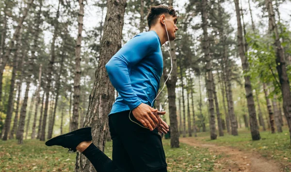 Изображение молодого человека, бегущего утром на свежем воздухе на фоне леса. Мужчина занимался в парке и слушал музыку в наушниках. Люди и спорт — стоковое фото