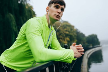 Sisli göl arka planında yapılan çalışmadan sonra sabah dinlenen genç atletik adamın yatay yakın plan portresi. Sporcu erkek parkta egzersiz yapıyor ve kulaklıkla müzik dinliyor..